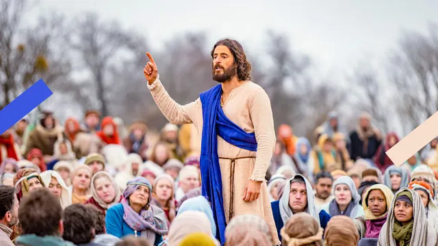 Je ziet een beeld uit de serie The Chosen. Jonathan Roumie, de acteur die Jezus speelt, spreekt een grote groep mensen toe. Hij heeft een beige tuniek aan en een blauwe doek om zijn bovenlichaam. Hij kijkt geconcentreerd.