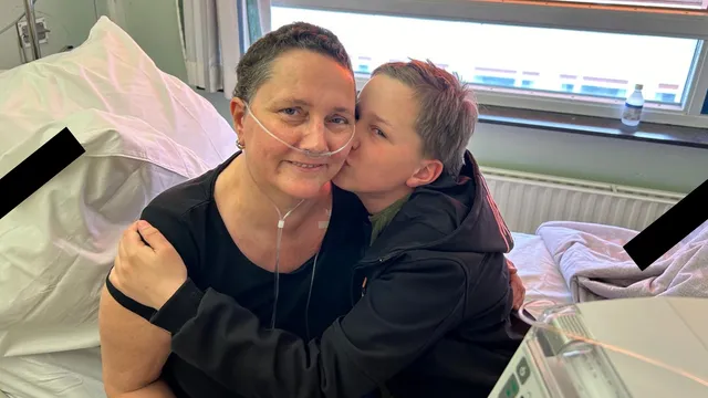 Dit is een foto waarop een jongen (Levi) een kus geeft op de wang van zijn moeder. Ze heeft kort haar en er loopt een slangetje naar haar neus. Ze zitten samen op een ziekenhuisbed.