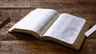 Je ziet een opengeslagen Bijbel liggen op een houten tafel.