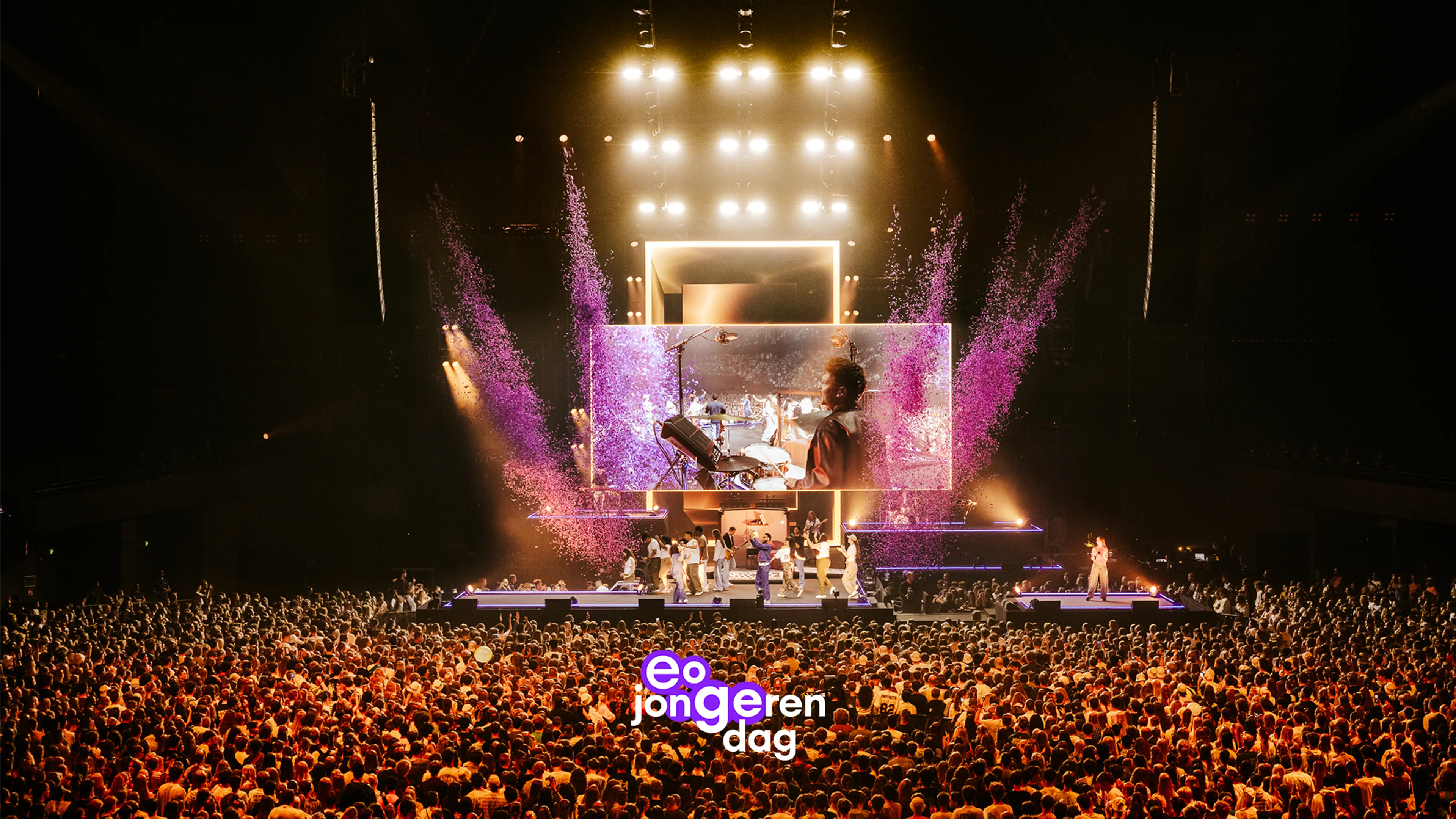 eo jongerendag is een groots indoor concertmoment in een arena, met een dicht opeengepakte menigte voor het podium en een centrale opstelling van lichtframes en een LED-videowand waarop een muzikant met toetsen zichtbaar is, terwijl het publiek en de band op de achtergrond te zien zijn. Paarse confetti- of sliertfonteinen en paars-accenten dragen bij aan de feestelijke sfeer rond het podium.