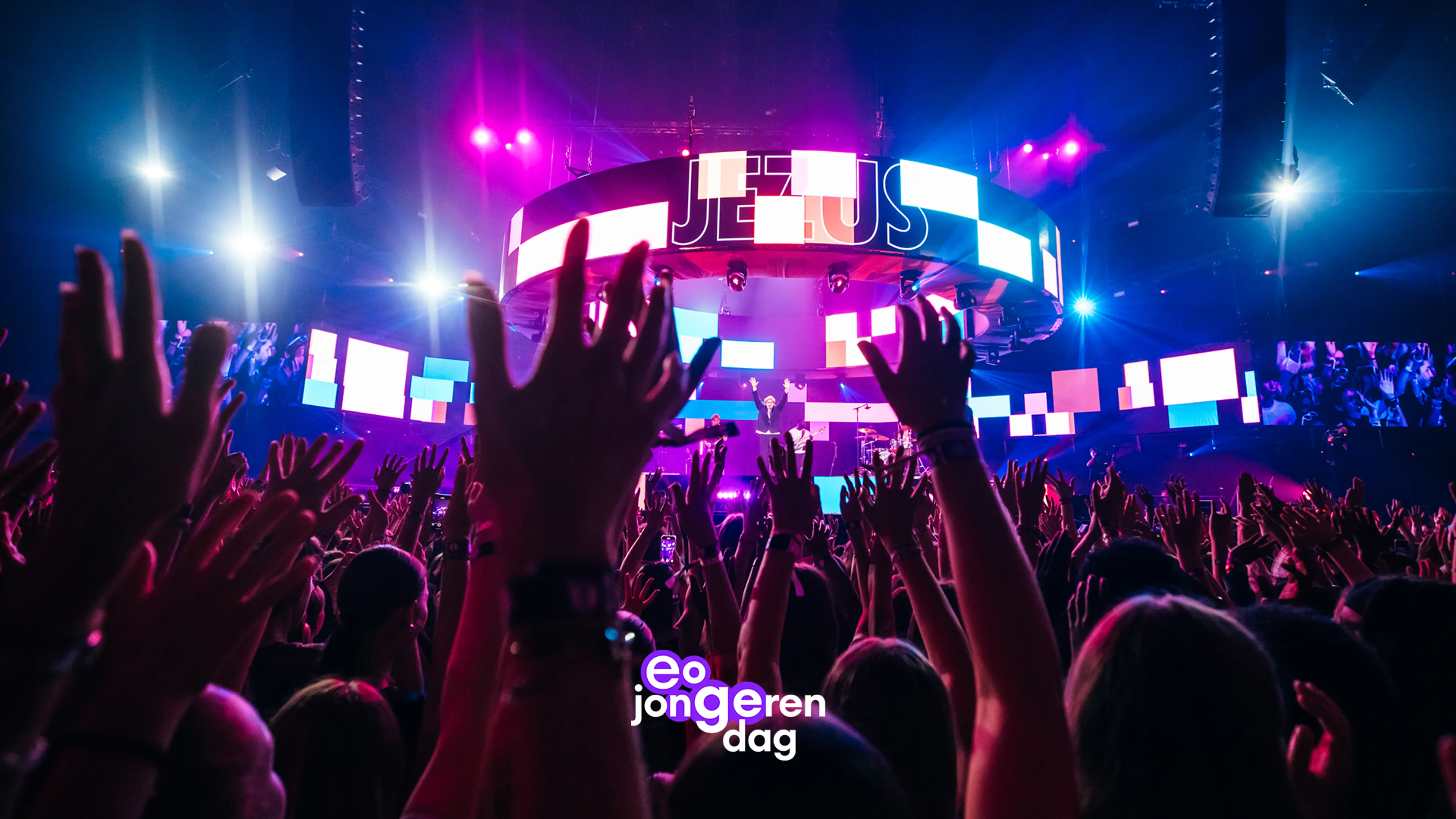EO Jongerendag is een christelijk jongeren-evenement met een concert- of worshipkarakter, zoals aangekondigd door het witte logo onderaan het podium. Het beeld toont een grote, drukbezochte indoor bijeenkomst met een viervoudig verlichte podium en een massale menigte die handen omhoog steekt richting het podium terwijl het woord “JEZUS” op een ronde LED-ring verschijnt.