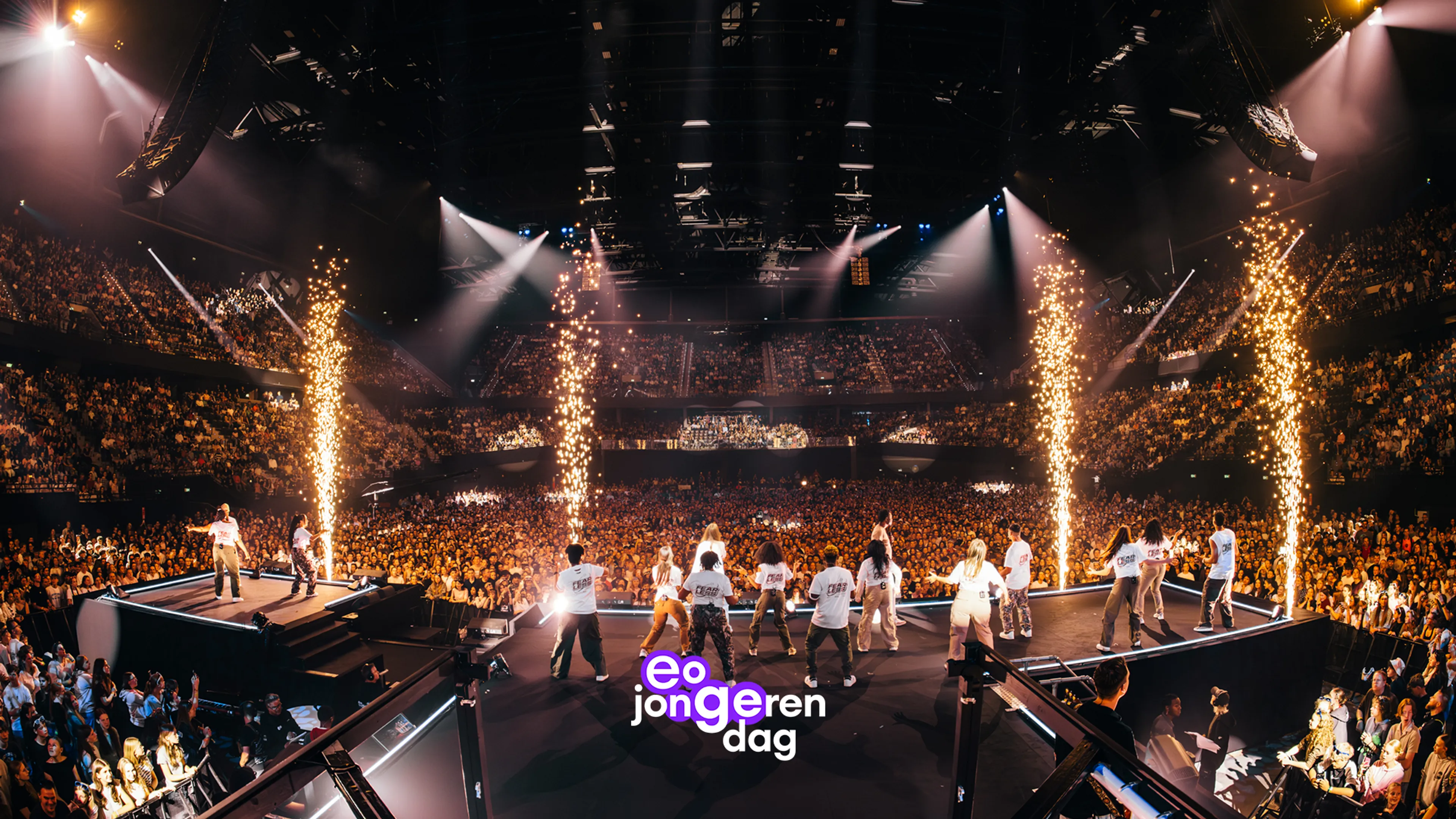 eo jongerendag is een grootschalige podiumshow waarbij een groep jonge performers in witte kleding in formatie naar het publiek beweegt, omlijnd door verticale pyrotechniek en fel verlichte rooklucht, met een duidelijk brandinglogo onderaan. De scène vindt plaats in een enorme indoor arena met tribunes, een verhoogd zijpodium en veel audiovisuele elementen die de feestelijke show benadrukken.