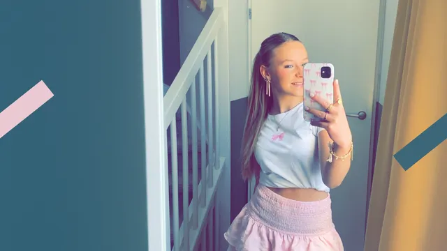 Je ziet hier Lenthe, die een foto maakt in de spiegel van een gang. Ze draagt een roze rokje en een wit t-shirt en lacht in de camera.