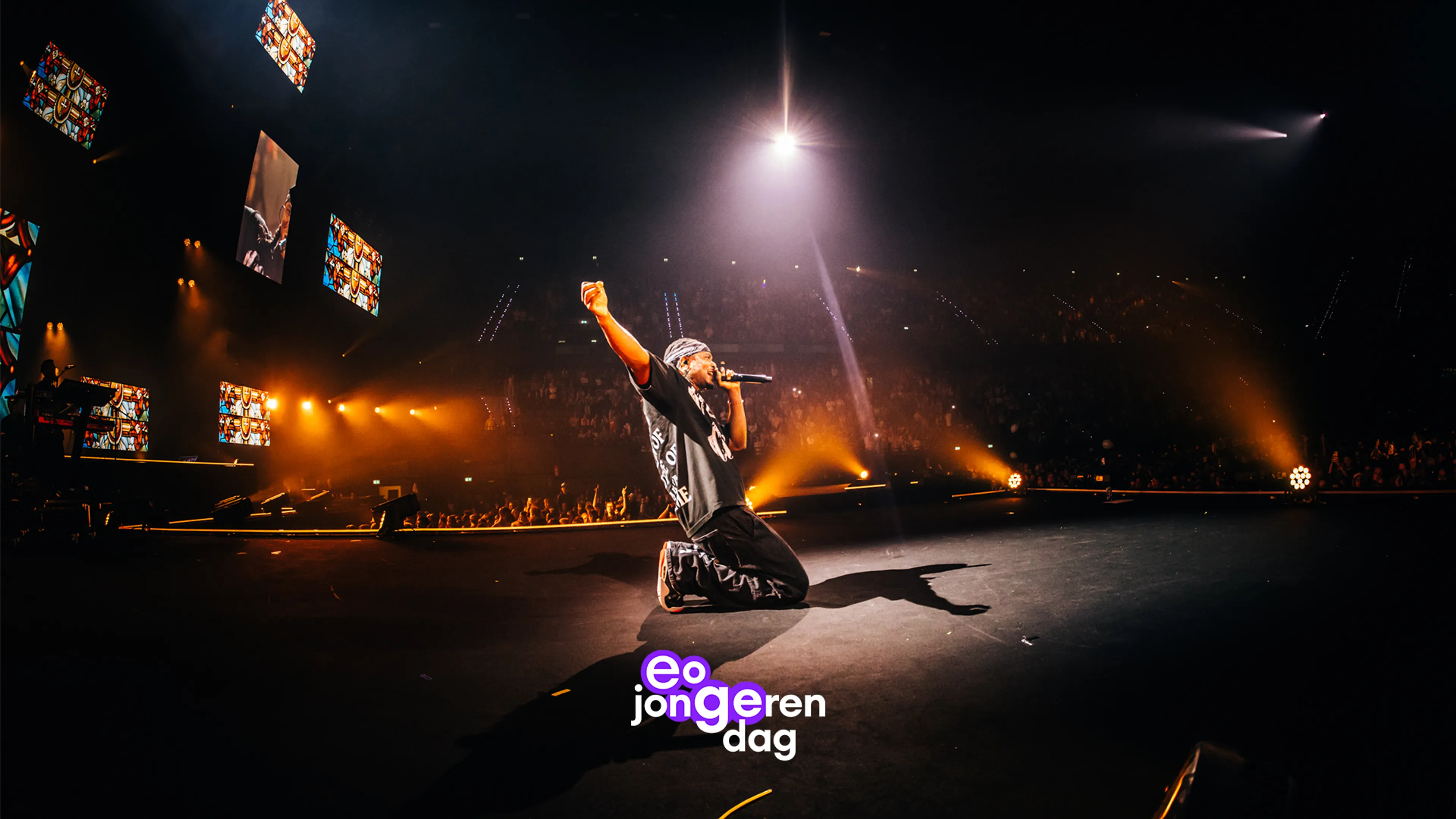 EO Jongerendag: Een concertachtige scène op een donker podium in een overdekte arena, waar een jonge mannelijke artiest in zwarte kleding knielt en met één arm een vuist omhoog steekt terwijl hij een microfoon vasthoudt; sterke witte en warme oranje belichting snijden door rook en benadrukken het moment, met een tribune vol toeschouwers en kleurrijke schermen op de achtergrond.