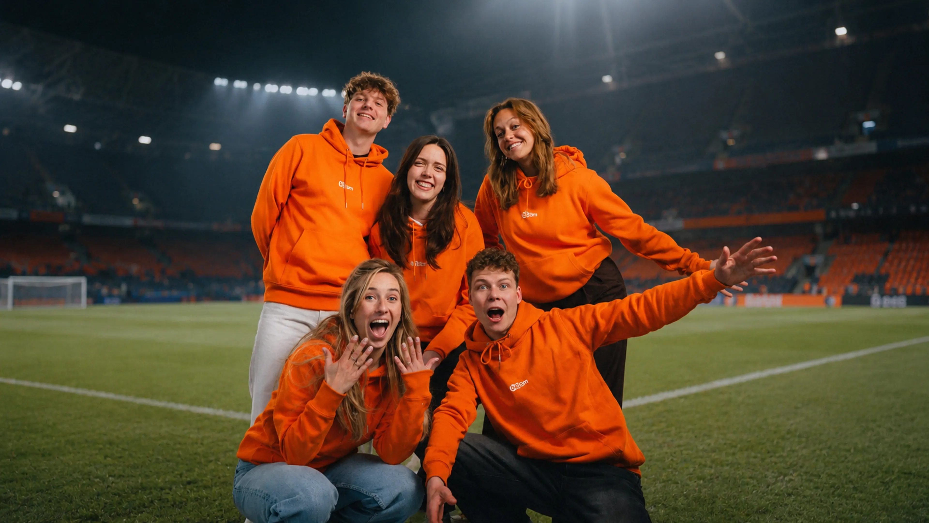 Fornamen? The primary subject is een groep van vijf jonge volwassenen op een voetbalveld in een stadion, allemaal in feloranje hoodies met een klein wit logo. Ze poseren feestelijk voor de camera, met twee vooraan knielend zichtbaar als ze juichend reageren, terwijl de drie achter hen ontspannen glimlachen.