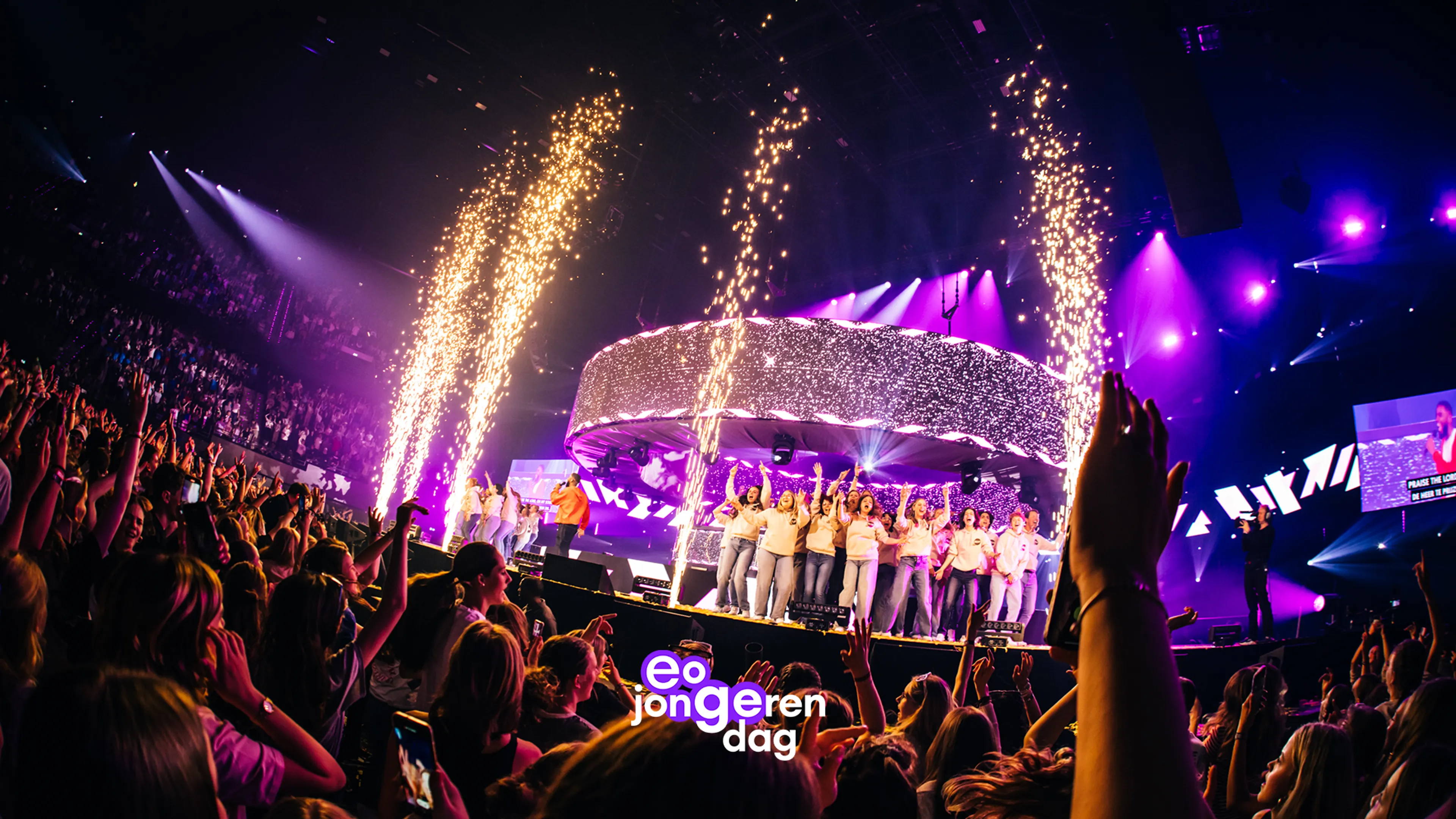 EO Jongerendag: Een overvolle indoorarena toont een feestelijke scène rondom een groot podium, waar een groep jonge performers dicht bij elkaar staat terwijl een spreker of zanger in oranje outfit naar het publiek kijkt en een groot videoscherm een close-up toont; de scène wordt verlicht door paars-roze verlichting en glitterende LED-constructies met gouden fonteinen, terwijl een juichende menigte het podium en tribunes vult en het logo “eo jongerendag” zichtbaar is.