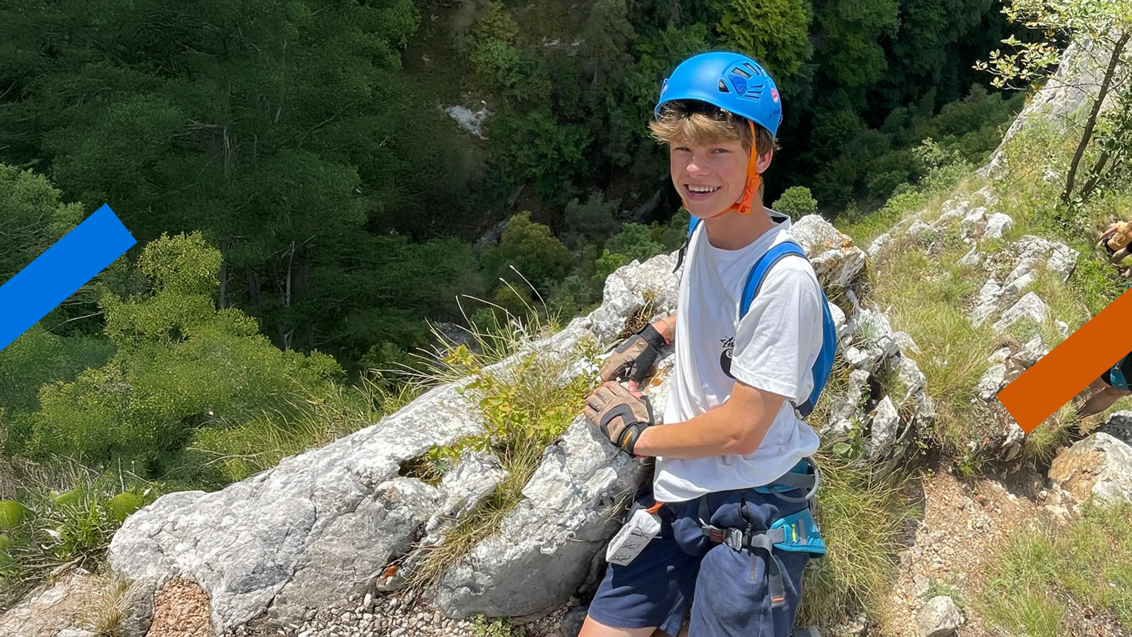 Je ziet een jongen in blauwe klimuitrusting op een berg bij een rots staan, dit is Jesse. Hij lacht naar de camera.