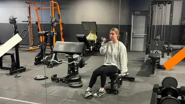 Je ziet een sportschool, waar Femke op een bankje zit. Ze houdt een dumbbell in haar hand.
