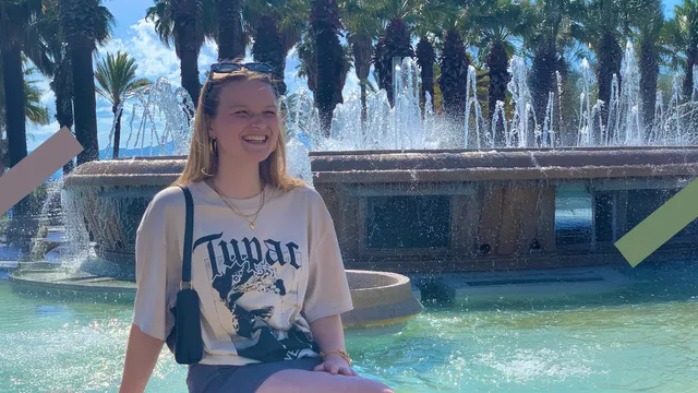 Je ziet een meisje lachend voor een fontein zitten. Dit is Anne Eva. De zon schijnt en ze draagt zomerkleding.