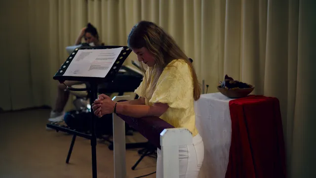 Rosa knielt op een bankje terwijl ze belijdenis doet in een kerk. Ze heeft een geel shirt aan en haar handen zijn gevouwen.