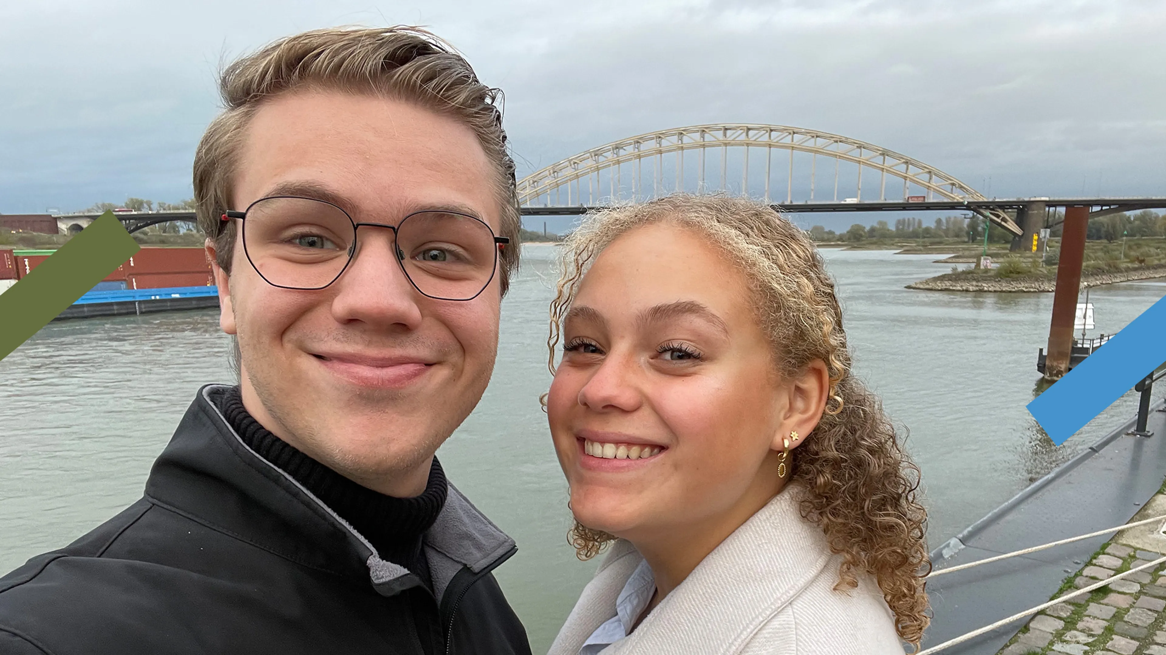 Een selfie met een jongen en een meisje voor een rivier. Op de achtergrond is ook een brug te zien en een boot. De jongen heeft een zwarte trui aan en het meisje een witte jas.