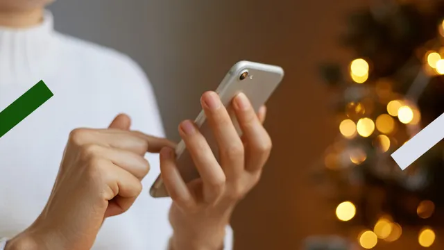 Je ziet de schouders en handen van een vrouw. Zij heeft een witte telefoon in haar hand. Je kunt het scherm niet zien. Rechts op de achtergrond staat een boom, waarvan je vooral de lichtjes ziet.