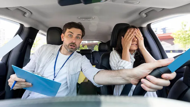 Een geschrokken rijleraar pakt het stuur over van een meisje dat rijles heeft en beide handen voor haar ogen heeft geslagen.