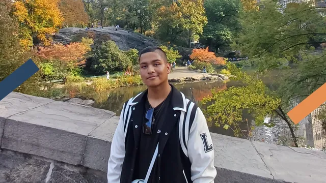 Een jongen, met een wit en zwarte jas aan, staat voor een muurtje. Erachter is een meertje te zien met bomen eromheen met rode en groene bladeren.