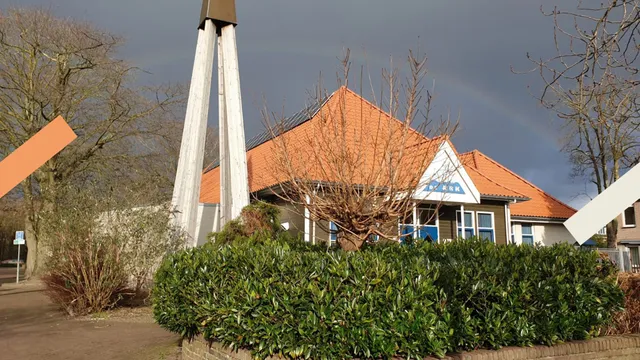 Een regenboog is te zien in de lucht. Er staat een kerk met een oranje dakpannen. Een gedeelte van de kerk is niet te zien door bosjes.