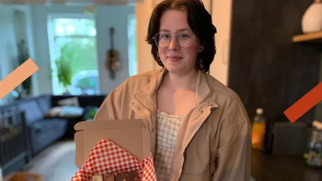 Een meisje met kort donkerbruin haar een beige jasje aan staat in een woonkamer. In haar handen heeft ze een kartonnen doosje met rood-wit geblokt inpakpapier met daarin stukjes fudge.