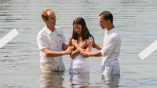 Meisje staat in het water met haar armen gekruist voor haar borstkas in het wit gekleed. Twee mannen hebben haar vast bij haar armen om haar zo te dopen.