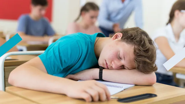 Een jongen met een blauw shirt ligt slapend op z'n tafel op school.