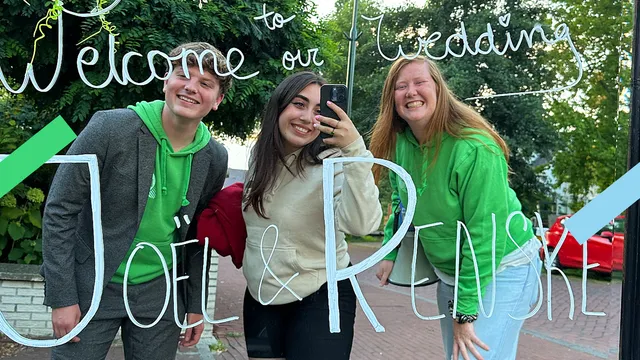 Redacteurs Gerben, C茅line en Wilke maken breed lachend een selfie in een spiegel waar 'welcome to our wedding' op staat met de namen van het bruidspaar: Jo毛l en Renske.