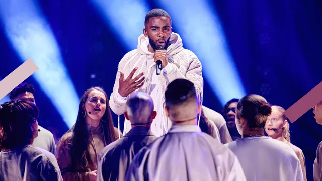 Je ziet Gideon Luciana op het podium staan met een microfoon in zijn hand. Hij heeft witte kleding aan en er staat een gospelkoor met witte kleding om hen heen.