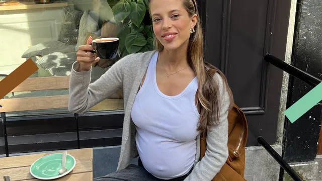 Zara zit hoogzwanger op een terrasje en toont trots haar kopje koffie. Ze draagt een wit shirt.