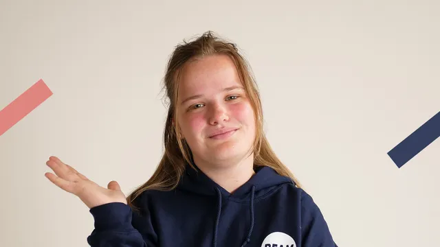 Een meisje met lang donkerblond haar en een donkerblauwe trui aan is te zien vanaf haar schouders. Ze houdt een hand vragend omhoog.