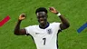 Voetballer Bukayo Saka loopt met het witte shirt van Engeland juichend over het voetbalveld. Hij houdt beide handen in de lucht en kijkt blij.